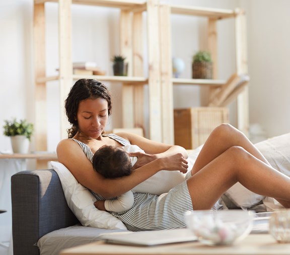 Serious calm careful young black mom sitting on sofa bed and breastfeeding baby while holding son in arms